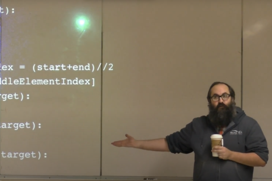 A bearded man standing in front of a white board with code projected