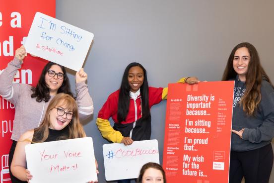 Five women holding up signs with #SitWithMe messages, posing while standing and sitting in the RedChair