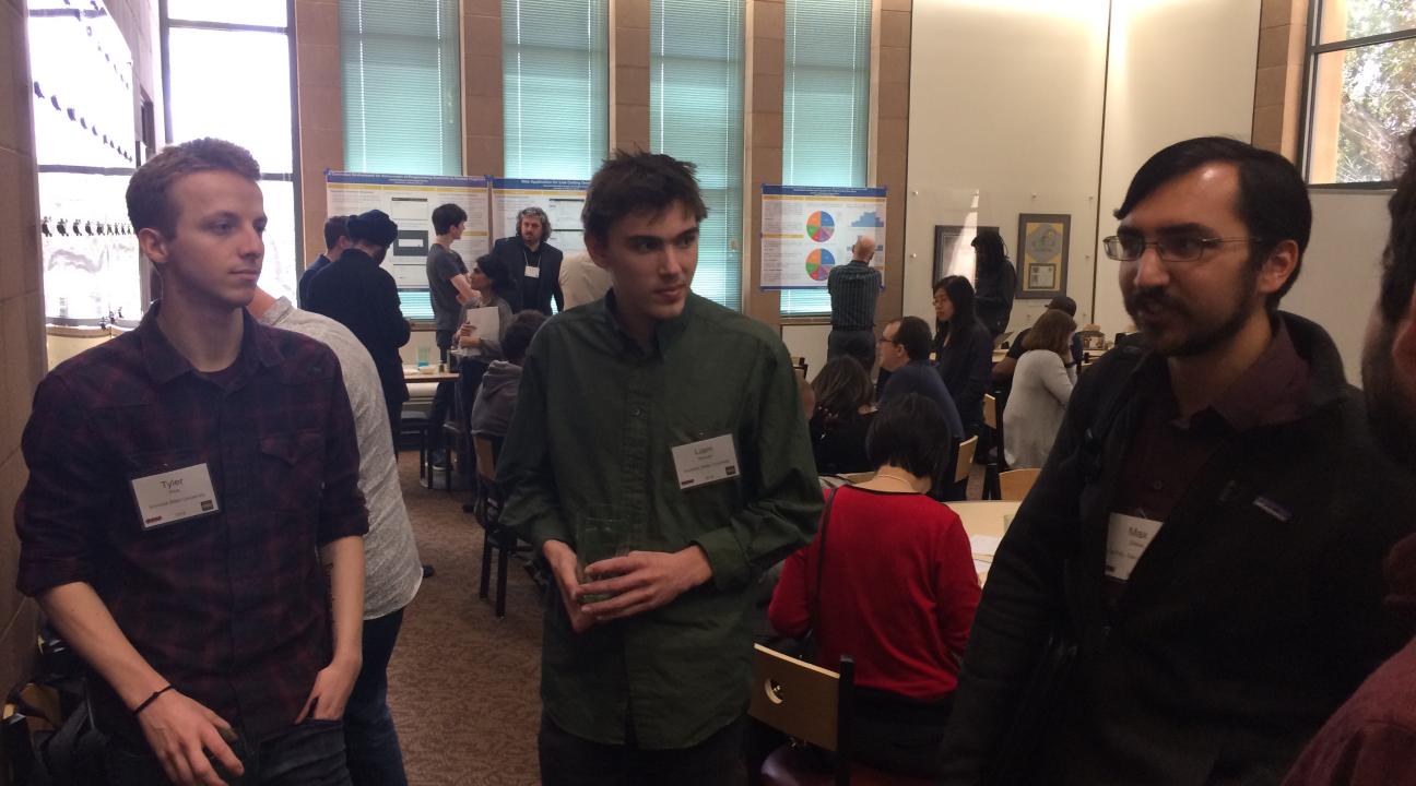 A student looks at a poster, not visible, while three students face him in discussion about the work