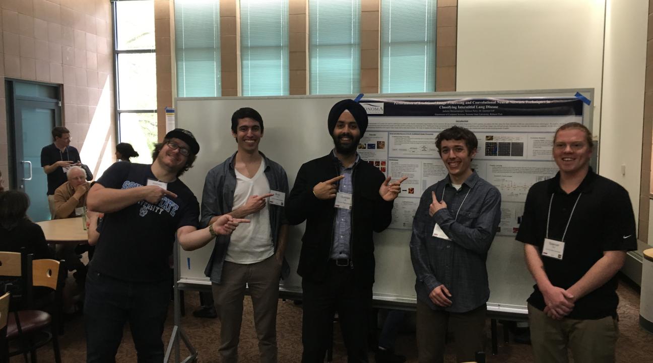 Four students and a professor pose in front of a poster in a middle of a room, pointing backwards to it, smiling