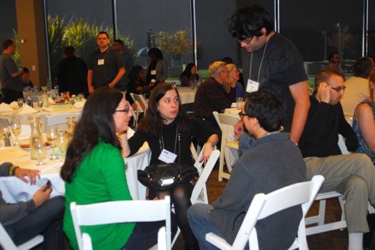 Three people sitting at a table in a crowded room, talking excitedly