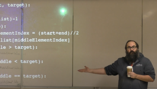 A bearded man standing in front of a white board with code projected