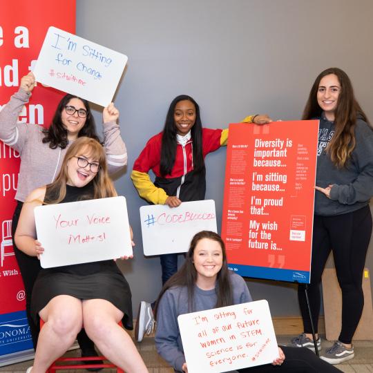 Five women holding up signs with #SitWithMe messages, posing while standing and sitting in the RedChair