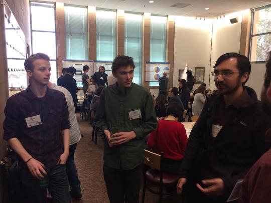 A student looks at a poster, not visible, while three students face him in discussion about the work