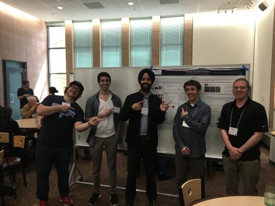 Four students and a professor pose in front of a poster in a middle of a room, pointing backwards to it, smiling